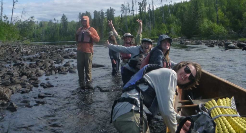 A group of students stand in ankle-deep water of a river near a canoe. There are green trees in the background. 